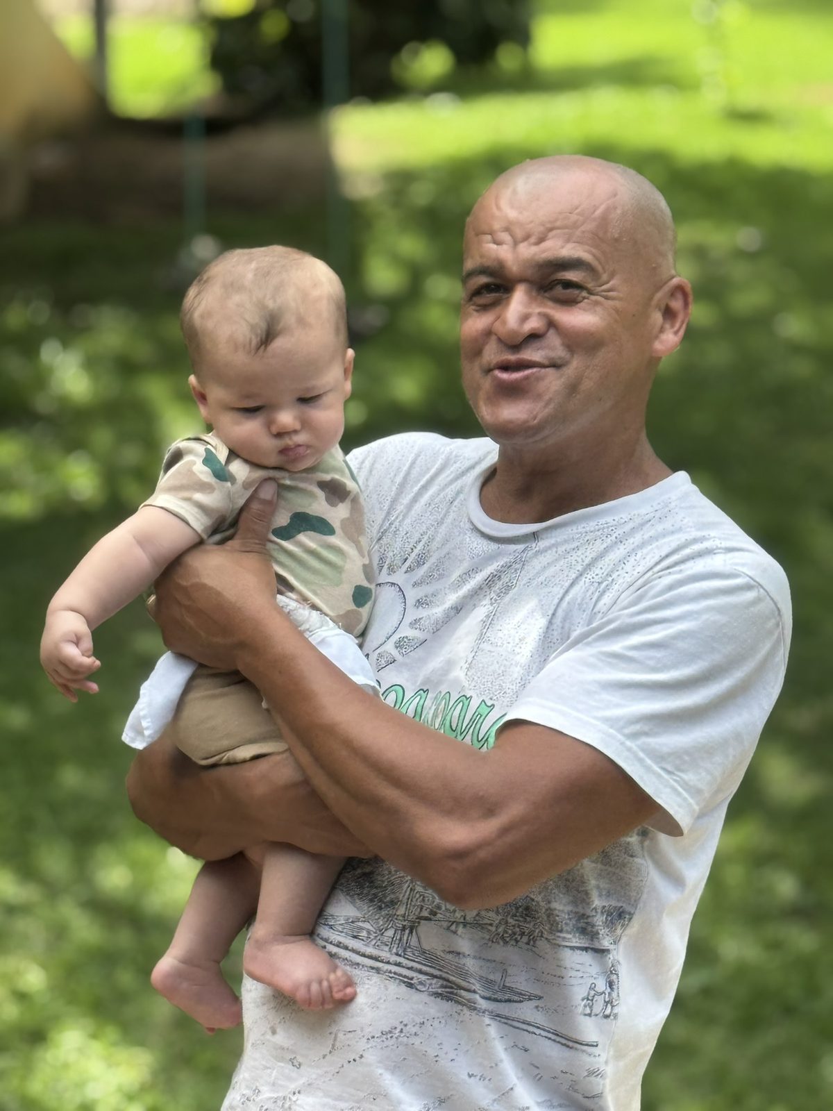 Avô sorrindo com o Benjamin no colo, no jardim.