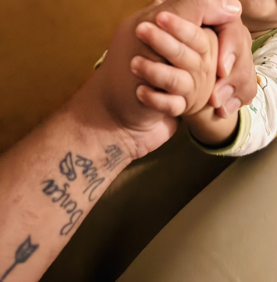 O braço tatuado do pai segura as mãozinhas do Benjamin.